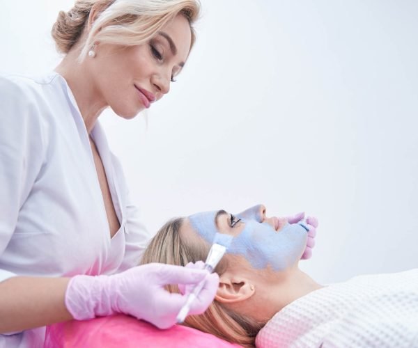 Cosmetician spreading blue clay paste over facial skin of client using two brushes